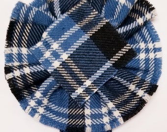 Puede incluir: Una roseta de tela tartán azul, blanco y negro con bordes deshilachados. La roseta está atada con dos tiras largas de tela. El texto "Clark, Ancient" está impreso debajo de la roseta.