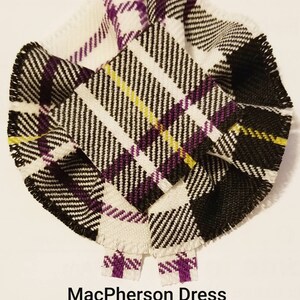 Può includere: Un elemento di tessuto circolare con un motivo tartan MacPherson Dress Ancient. Il design presenta una base bianca con strisce nere, viola e gialle. I bordi sono sfilacciati e il testo "MacPherson Dress Ancient" è visibile.