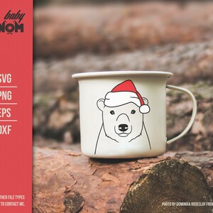 May include: A white enamel mug featuring a polar bear wearing a red Santa hat. The mug has a black outline drawing of the bear's face. The mug is on a wooden surface. The text "baby GNOM" is at the top left.