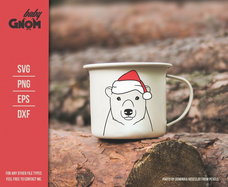 May include: A white enamel mug featuring a polar bear wearing a red Santa hat. The mug has a black outline drawing of the bear's face. The mug is on a wooden surface. The text "baby GNOM" is at the top left.