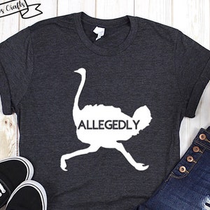 May include: A dark gray t-shirt with a white silhouette of an ostrich and the word "ALLEGEDLY" printed on it. The shirt is shown on a white wooden background with a pair of black Vans shoes and blue jeans.