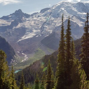 May include: A panoramic view of a snow-capped mountain peak with a valley in the foreground. The valley is covered in lush green vegetation and a winding river. The mountain is surrounded by a dense forest of evergreen trees.