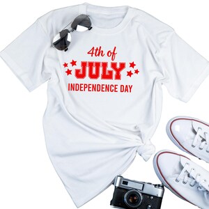 Könnte beinhalten: Weißes T-Shirt mit dem roten Text "4th of July Independence Day". Das Shirt ist mit Sonnenbrille, weißen Turnschuhen und einer Vintage-Kamera gestylt. Das Shirt ist an der Taille geknotet.
