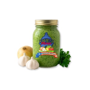May include: A glass jar filled with green sofrito, labeled "Sofrito Maria" with a blue, red, and white design. The jar is topped with a gold lid and surrounded by fresh garlic, an onion, and parsley. The label reads "homemade-with-love."