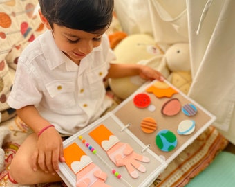May include: A child is sitting on a bed and looking at a felt activity book with colorful felt shapes and beads. The book has a white cover with a light gray border.