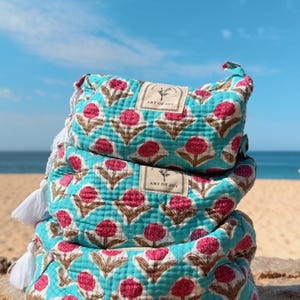 May include: A stack of three quilted pouches in a bright turquoise fabric featuring a repeating pattern of pink flowers with brown stems. Each pouch has a white tassel and a rectangular label with the text Art of Joy and a flower icon.
