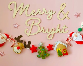 May include: A felt Christmas garland with Santa, reindeer, a gingerbread house, a Christmas tree, candy canes, and stars. The garland is strung on a white string and has the words "Merry & Bright" written in wood above it.
