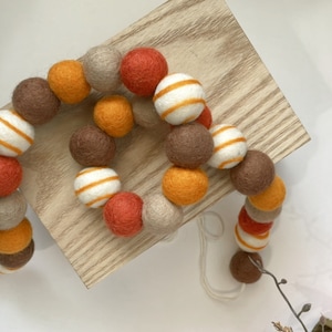 May include: A garland of felt balls in orange, brown, beige, and white with stripes. The garland is made of wool and is perfect for fall decor.