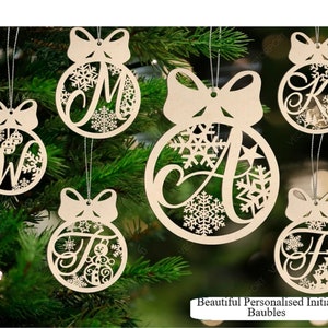 May include: A selection of wooden Christmas baubles, each featuring a different initial letter and snowflake patterns. The baubles are round with a bow detail and a silver string for hanging. The letters visible are W, M, A, K, and F. Text: "Beautiful Personalised Initial Baubles."