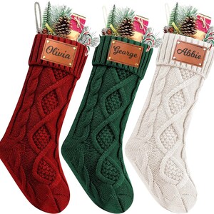 May include: Three cable-knit Christmas stockings in red, green, and cream. Each stocking has a personalized leather patch with a name: Olivia, George, and Abbie. Stockings are filled with gifts, candy canes, and pine sprigs.