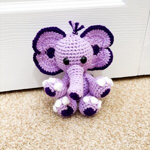 May include: A handmade crochet elephant toy in shades of purple and white. The elephant has large, floppy ears with dark purple trim, small black eyes, and white accents on its feet. The toy is sitting on a beige carpet.