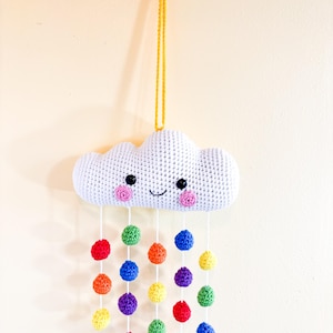 May include: A white crocheted cloud with a smiling face and pink cheeks hangs from a yellow cord. Below, colorful crocheted balls in red, orange, yellow, green, blue, and purple dangle, resembling raindrops. This handmade item is a decorative piece.