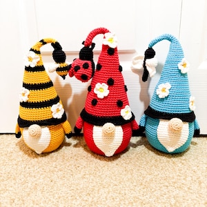 May include: Three crochet gnomes with whimsical designs. One gnome is yellow and black with a bee, another is red with black spots and a ladybug, and the third is blue with white wings. Each gnome has a white heart-shaped belly and small white flowers.