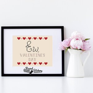May include: A framed cross-stitch design featuring the text "Ew, Valentine's Day" in black lettering on a beige background. The design is bordered by a row of red hearts.