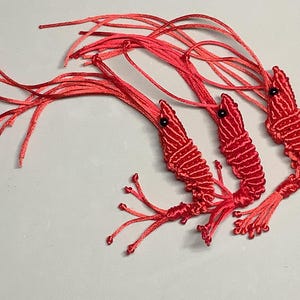 May include: Three handmade red shrimp ornaments with black eyes and detailed stitching. Each shrimp has long, red thread antennae and legs. The shrimp are arranged on a white surface.