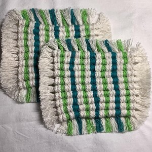 May include: Two square macrame coasters. Each coaster features a woven pattern of white, teal, and green stripes. The edges are finished with a fringe of white macrame. These coasters are designed to protect surfaces from drink condensation.