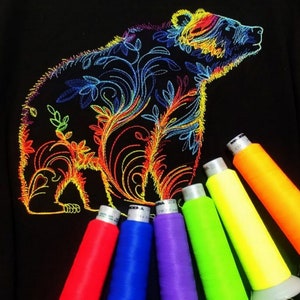 May include: A black t-shirt featuring an embroidered design of a bear with floral details. The bear is outlined in rainbow colours. The image also shows spools of neon coloured thread.
