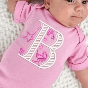 May include: A pink baby onesie with a white embroidered letter B, surrounded by stars, a crescent moon, and a baby carriage. The onesie has the text "Nata-Xa" above the letter B.