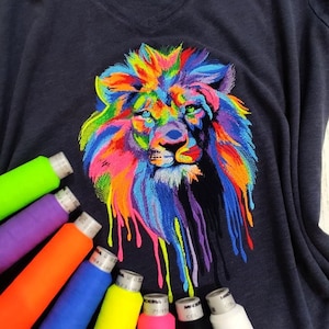 May include: A navy blue V-neck t-shirt with a vibrant lion design. The lion is depicted in a multitude of colours. Several spools of thread in various colours are arranged around the shirt, suggesting a crafting or sewing theme.