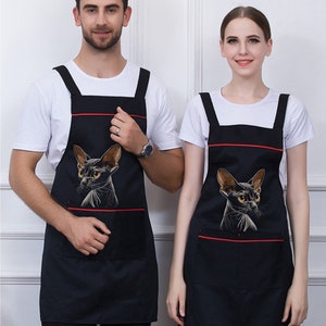 May include: Two black aprons with red trim and a black cat graphic. The aprons have a large pocket on the front.