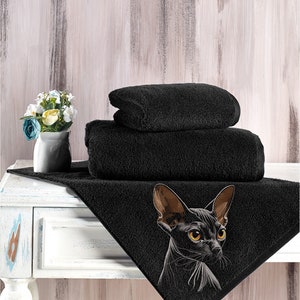 May include: A black bath towel with an embroidered image of a black Sphynx cat with yellow eyes. The towel is folded in thirds and placed on a white wooden surface.