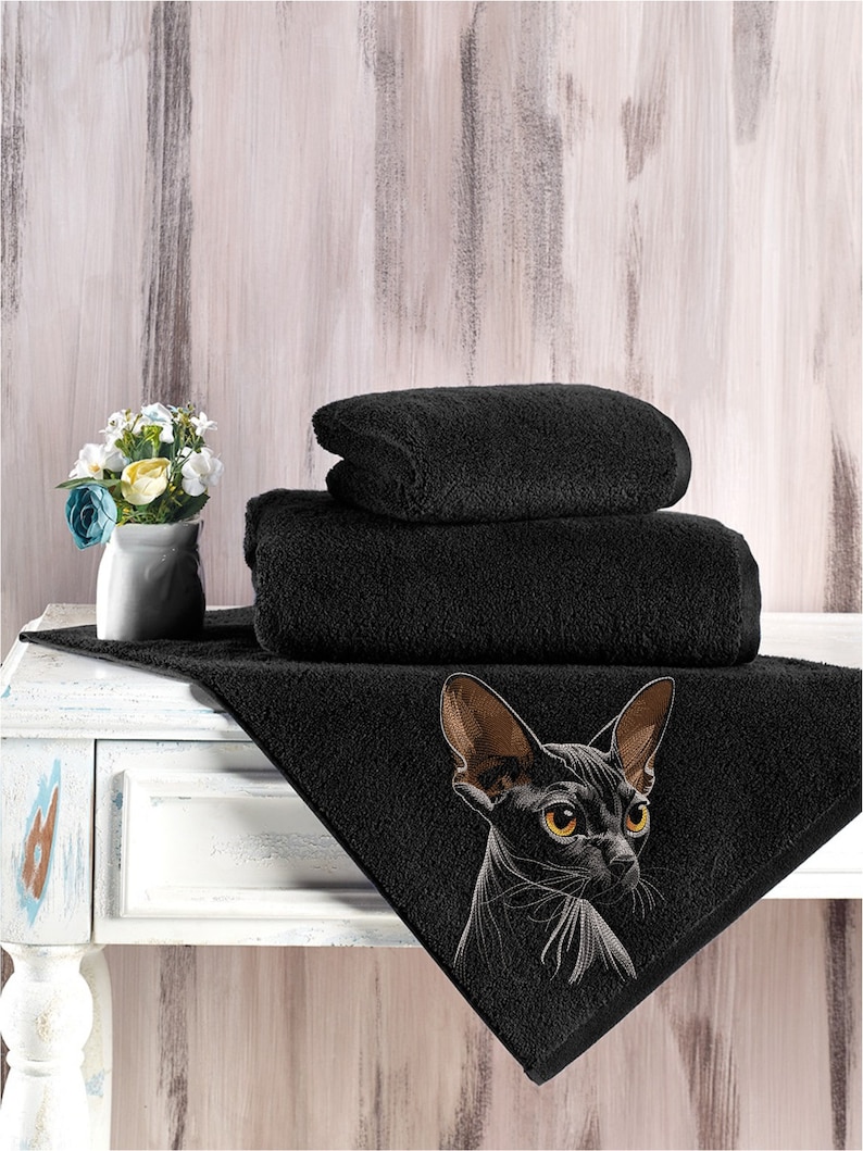 May include: A black bath towel with an embroidered image of a black Sphynx cat with yellow eyes. The towel is folded in thirds and placed on a white wooden surface.