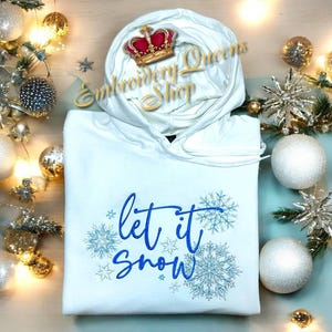 Puede incluir: Sudadera con capucha blanca con un diseño bordado en azul que dice "Let it snow" con copos de nieve alrededor del texto.