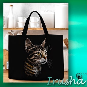 May include: Black tote bag with an embroidered image of a tabby cat with green eyes. The cat is looking to the right. The bag has two handles.