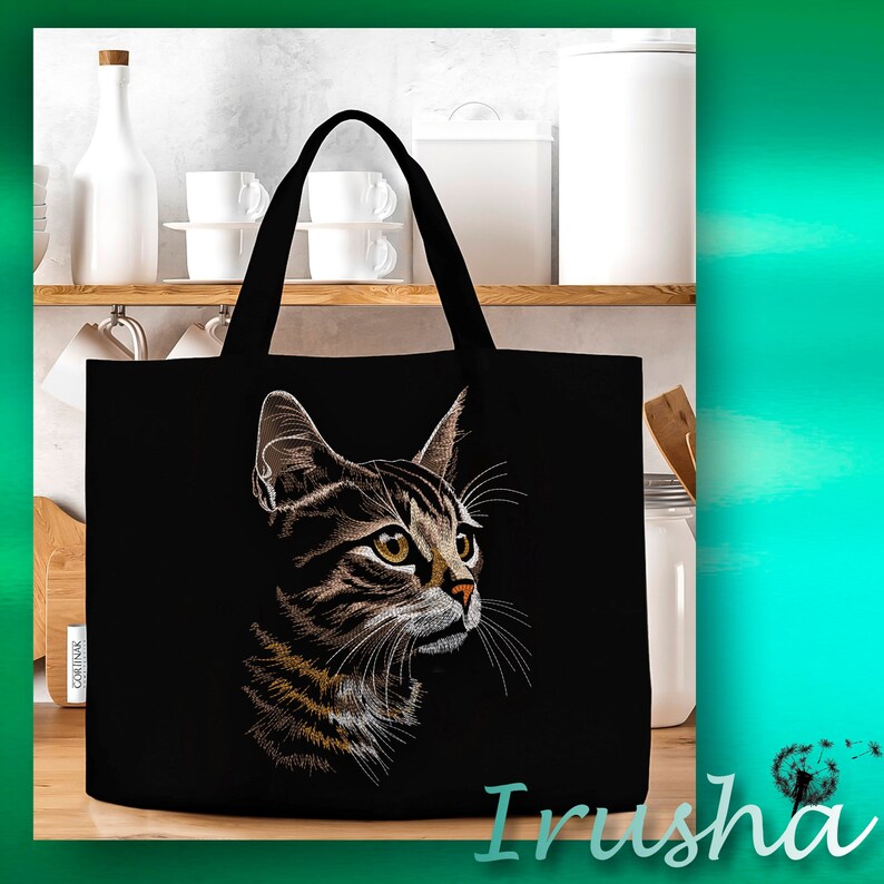 May include: Black tote bag with an embroidered image of a tabby cat with green eyes. The cat is looking to the right. The bag has two handles.