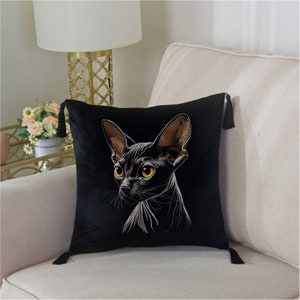 May include: Black velvet pillow with a white embroidered image of a hairless cat with orange eyes. The pillow has tassels on the corners.
