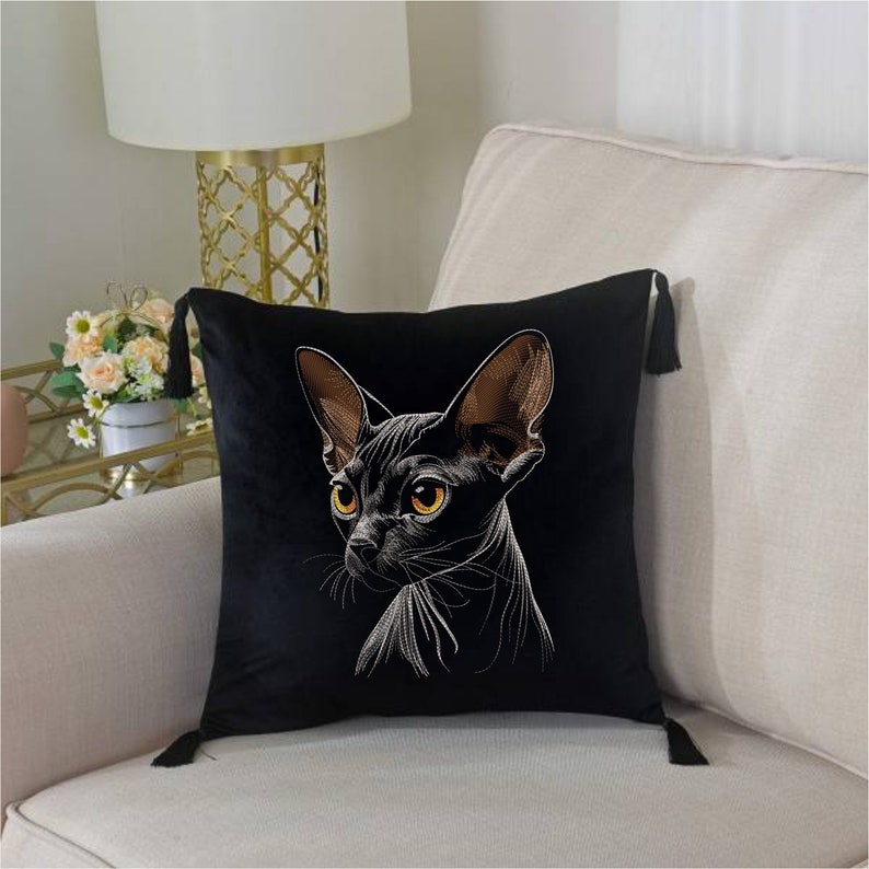 May include: Black velvet pillow with a white embroidered image of a hairless cat with orange eyes. The pillow has tassels on the corners.