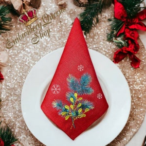May include: A red linen napkin with an embroidered design of a green and blue pine branch with yellow berries and white snowflakes. The napkin is folded into a triangle and placed on a white plate.