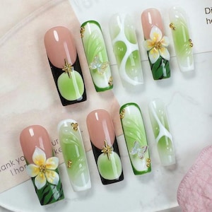 May include: A set of ten press-on nails featuring a variety of designs including green and white gradients, 3D white flower embellishments, translucent butterfly accents, gold metallic studs, and black circular patterns with gold star charms.