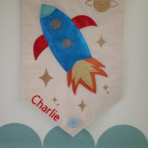 May include: A cream banner featuring a blue glitter rocket with red fins and gold detailing. The banner has the name "Charlie" in red, gold star accents, and a blue button. A gold glitter planet is at the top.