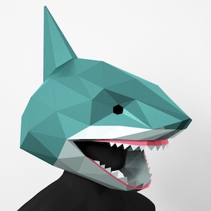 Puede incluir: Una máscara de tiburón de papel artesanal de color azul verdoso con una boca blanca y dientes rosados. La máscara tiene una aleta triangular en la parte superior y un ojo negro.