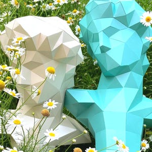 May include: Two geometric paper bust sculptures, one cream-colored and one teal, stand amidst a field of daisies. The sculptures have a modern, angular design. The daisies have white petals and yellow centers, adding a natural contrast to the sculptures.