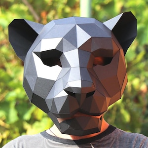 May include: A black geometric panther mask with triangular facets. The mask is worn by a person wearing a gray and white striped shirt, arms crossed. The background is blurred green foliage.