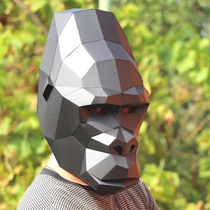 May include: A geometric, black gorilla mask with a polygonal design. The mask covers the entire head and face, with visible eye holes. The person wearing the mask is wearing a black and white striped shirt, with their arms crossed.