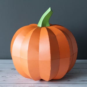 Könnte beinhalten: Ein dreidimensionaler Kürbis aus Papier mit einem leuchtend orangefarbenen Körper aus vertikalen Segmenten und einem hellgrünen Stiel. Der Kürbis hat eine glatte, matte Oberfläche und eine abgerundete, segmentierte Form mit Kanten.