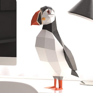 May include: A geometric papercraft puffin sculpture with a red beak and feet, standing on a white desk. The bird is black, white, and gray. A modern desk lamp with a gray shade and a white desk and chair are also in the image.