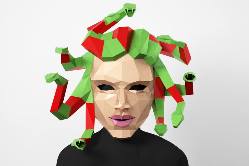 May include: A papercraft Medusa mask with green and red snakes. The mask has a geometric design with a light brown face, black eyes, and pink lips. The person is wearing a black top.