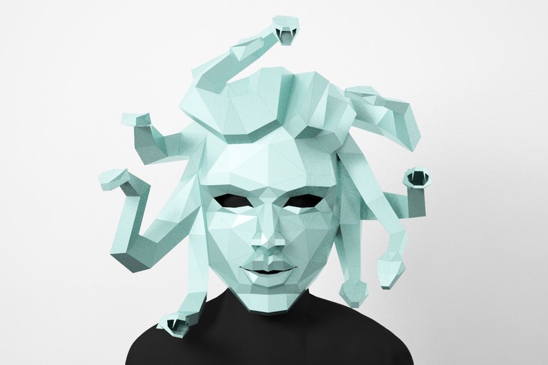 May include: A light blue geometric Medusa mask with snake-like hair. The mask has a polygonal design and is set against a white background. The person wearing the mask is wearing a black shirt.