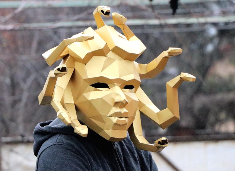 May include: A gold-colored Medusa mask with a geometric design. The mask features a detailed face with a prominent nose and mouth, and stylized snakes. The mask is worn over a dark gray hooded sweatshirt.