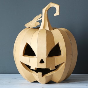 May include: A tan cardboard pumpkin sculpture featuring a carved jack-o'-lantern face with triangular eyes, a triangular nose, and a wide, jagged mouth. The pumpkin has a segmented, geometric construction with a curved stem and a single leaf.