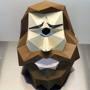 Può includere: Una scultura geometrica di carta di un volto barbuto. La scultura è principalmente marrone, con accenti bianchi, azzurri e neri. Il volto ha un aspetto stilizzato, con tratti angolari e un occhio chiuso.