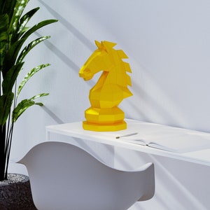 Puede incluir: Una pieza de ajedrez de caballero de origami amarilla se encuentra sobre un escritorio blanco con una silla blanca y una planta en maceta en el fondo.