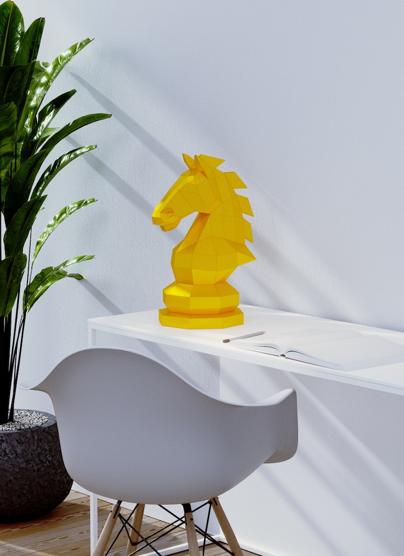 Puede incluir: Una pieza de ajedrez de caballero de origami amarilla se encuentra sobre un escritorio blanco con una silla blanca y una planta en maceta en el fondo.