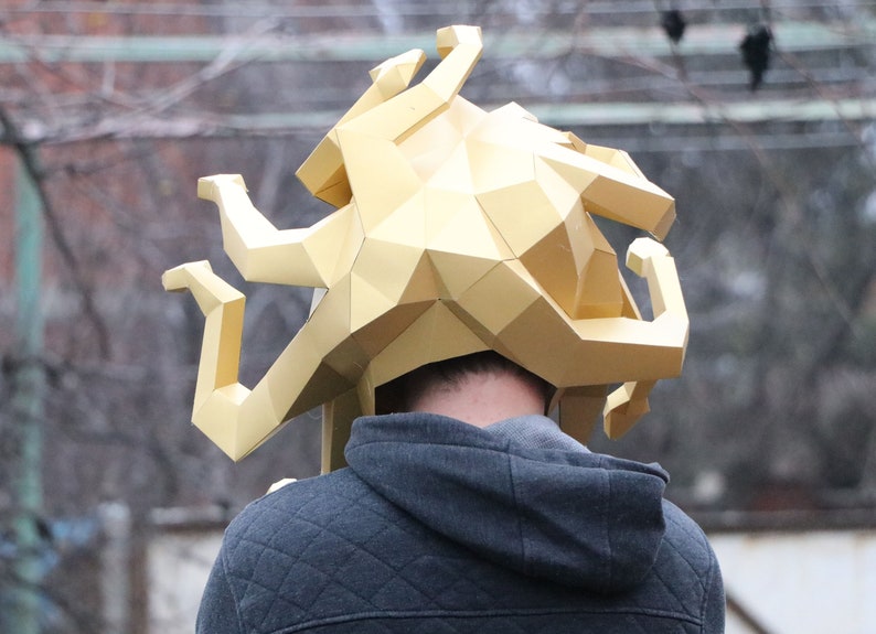 May include: A gold-colored, geometric-shaped headpiece, possibly a helmet or mask, is worn by a person. The headpiece has a polygonal design with multiple curved extensions. The person is wearing a gray hooded sweatshirt.