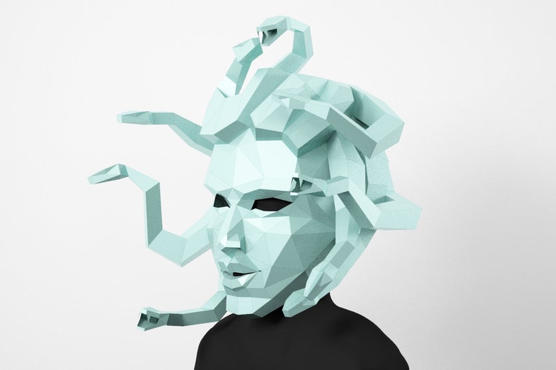 May include: A geometric, light blue Medusa mask with snake-like hair and arms. The mask is made of angular shapes and is set against a white background. The person is wearing a black top.