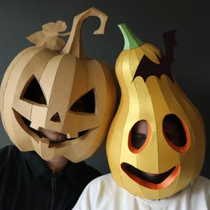 May include: Two papercraft masks shaped like pumpkins. The left mask is tan with a jagged, triangular-eyed jack-o'-lantern face and a stem. The right mask is yellow with a smooth, rounded face, orange eye and mouth cutouts, and a black bat silhouette.